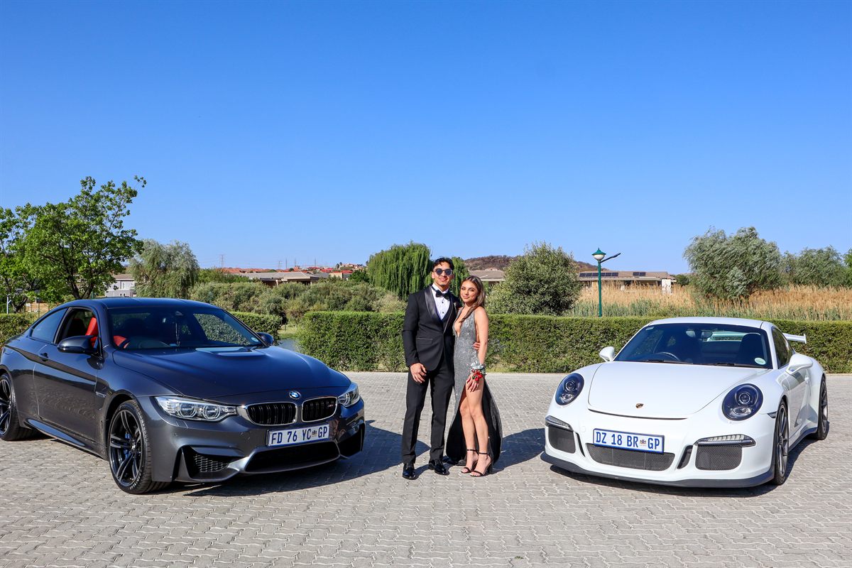 A couple in formal dress standing between a grey BMW M4 and a white Porsche 911