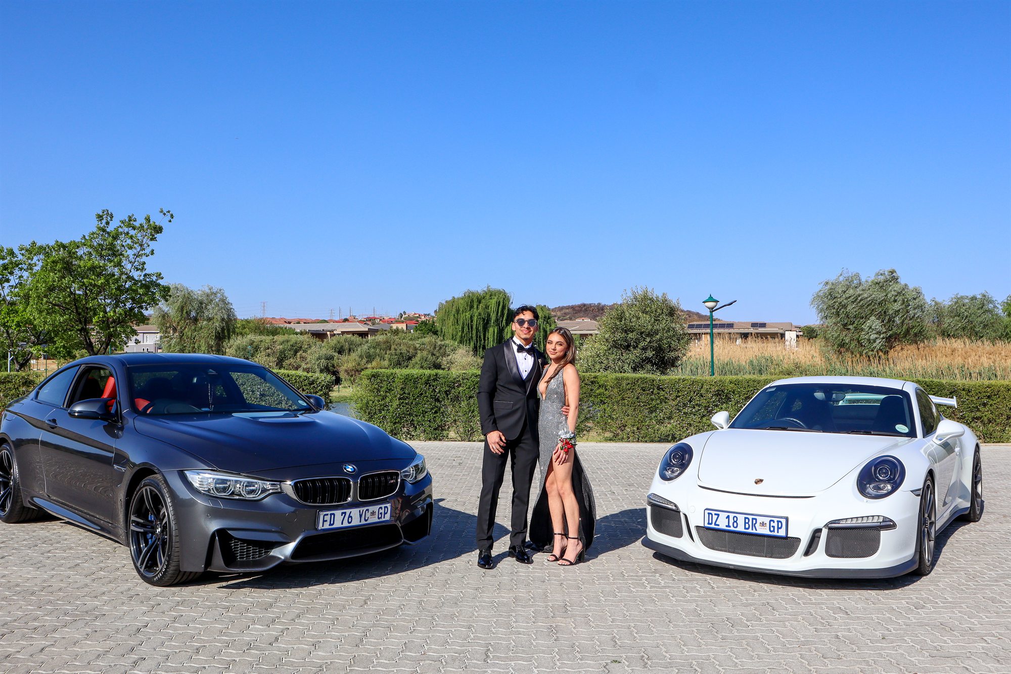 A couple in formal dress photographed between a BMW M4 and a Porsche 911 at a matric dance send-off