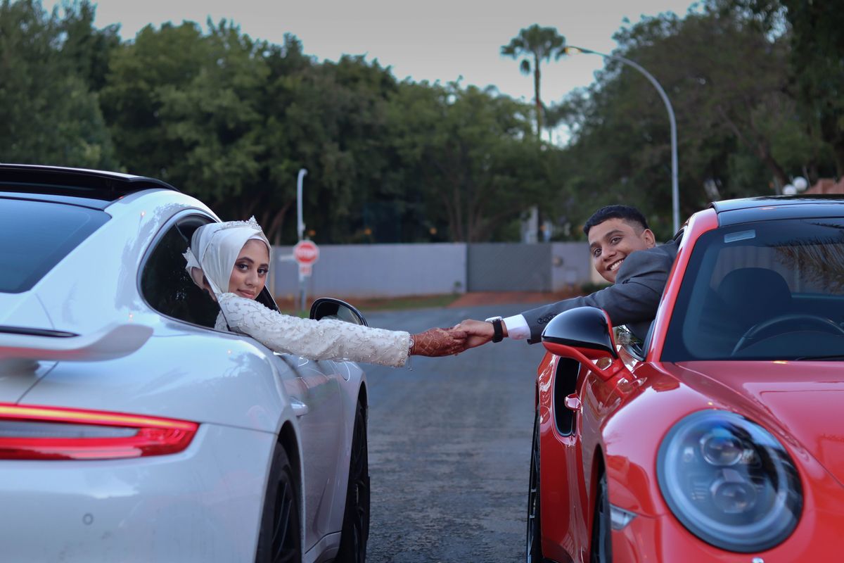 A bride and groom reaching for each other's hands through the windows of a white and a red Porsche