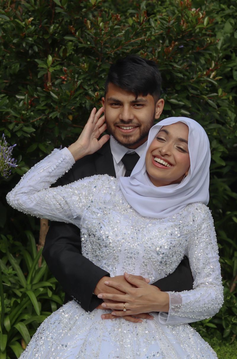 A groom holding his bride from behind, both laughing, greenery behind them