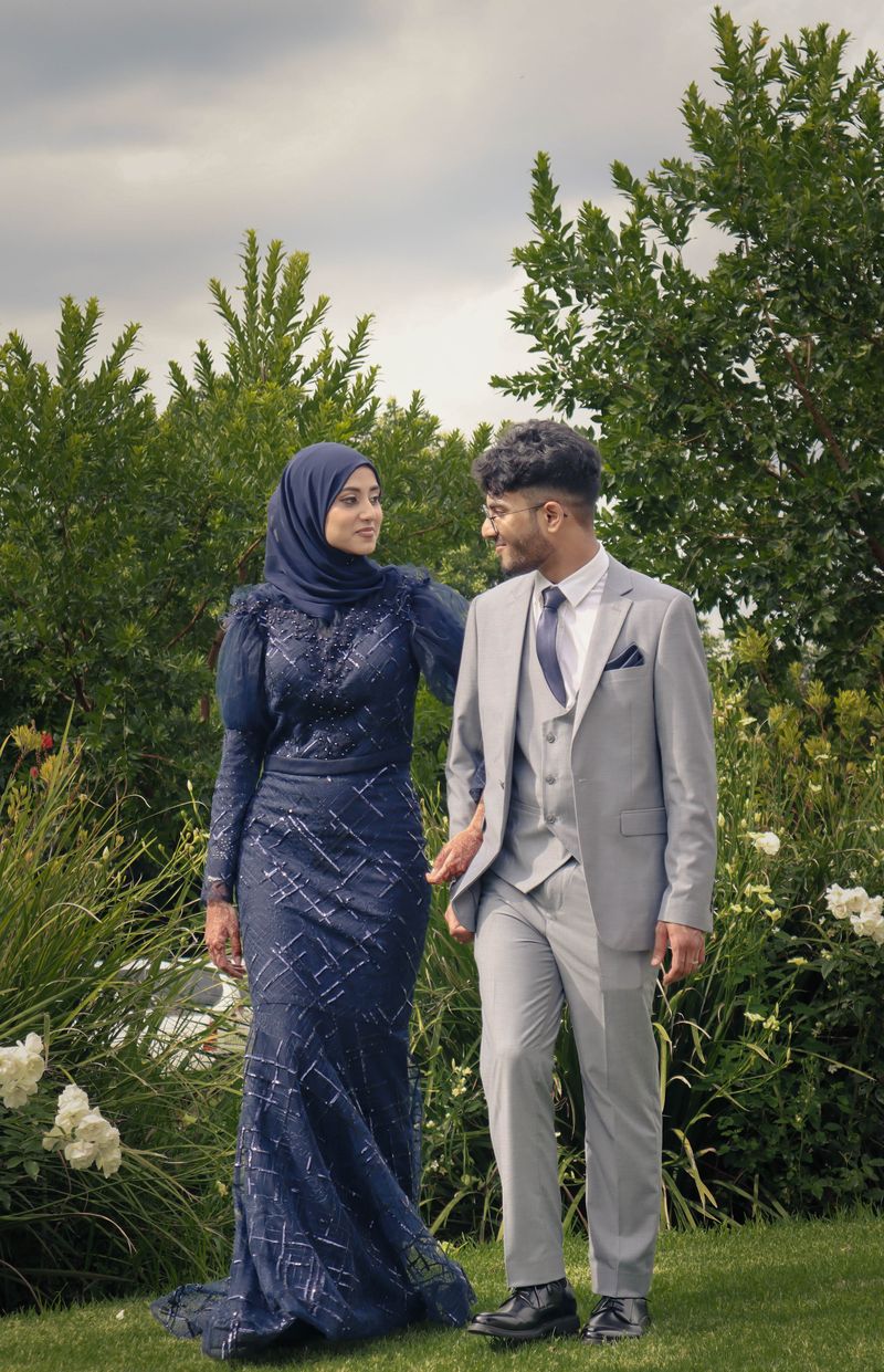 A couple walking through a garden at dusk, the bride in a navy beaded gown