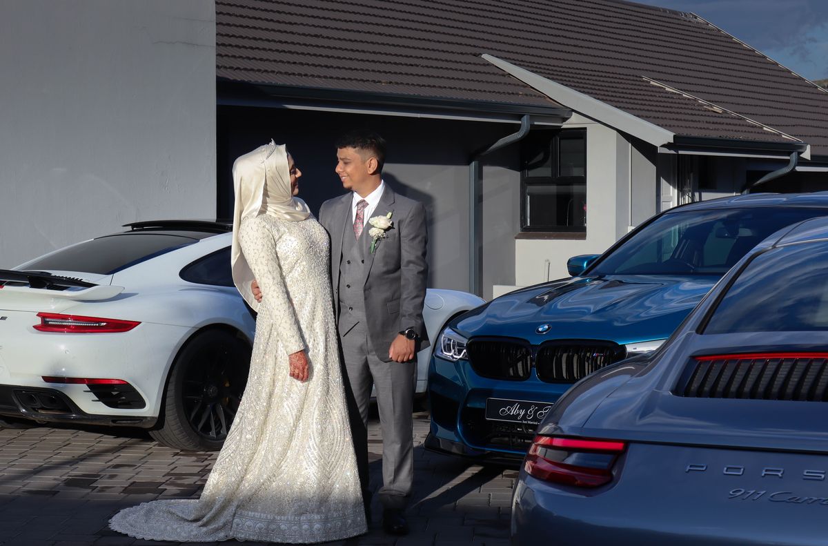 A bride and groom on a driveway flanked by a white Porsche, a blue BMW and a grey Porsche