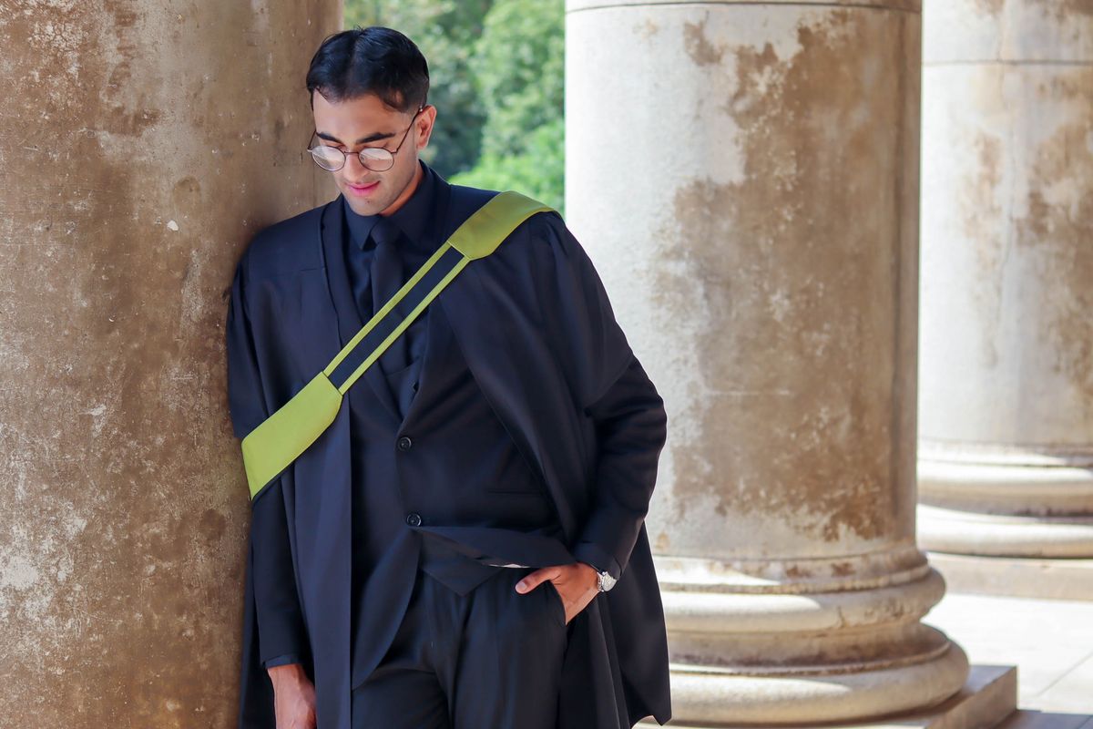 A graduate in cap and gown leaning against the stone columns of a university building