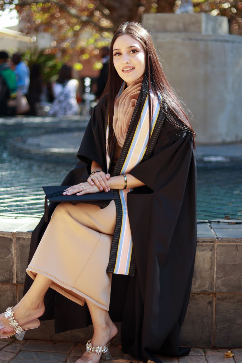 A graduate in cap and gown seated at the edge of a fountain, holding her cap