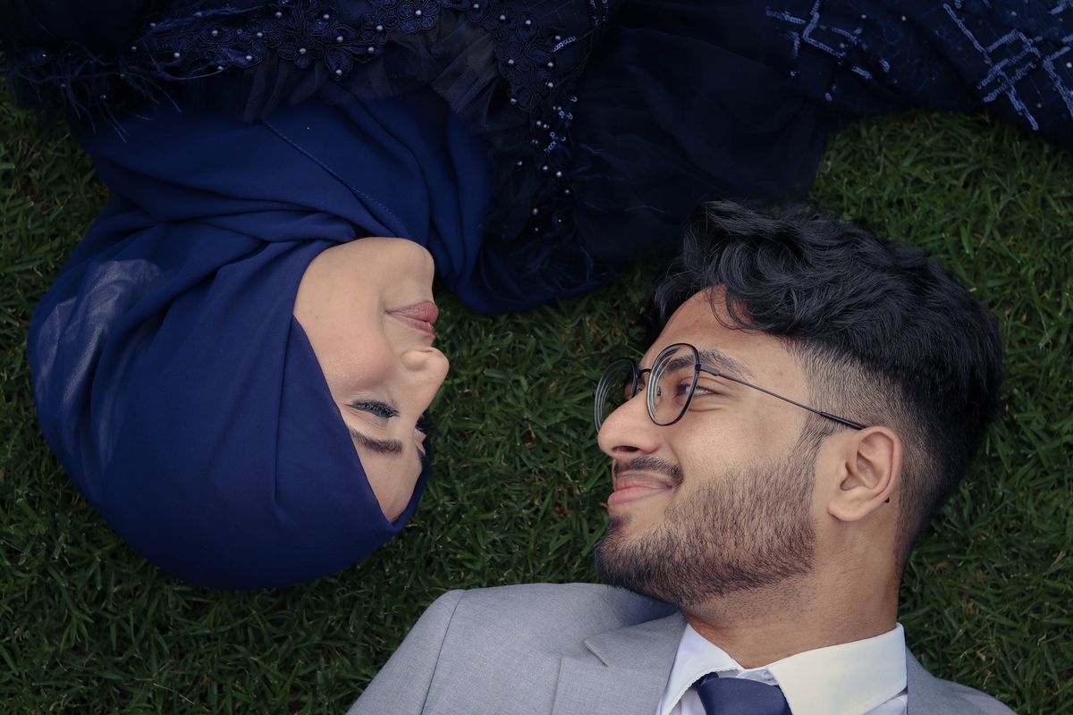 A couple lying head to head on grass, the bride in a navy gown and the groom in a light grey suit