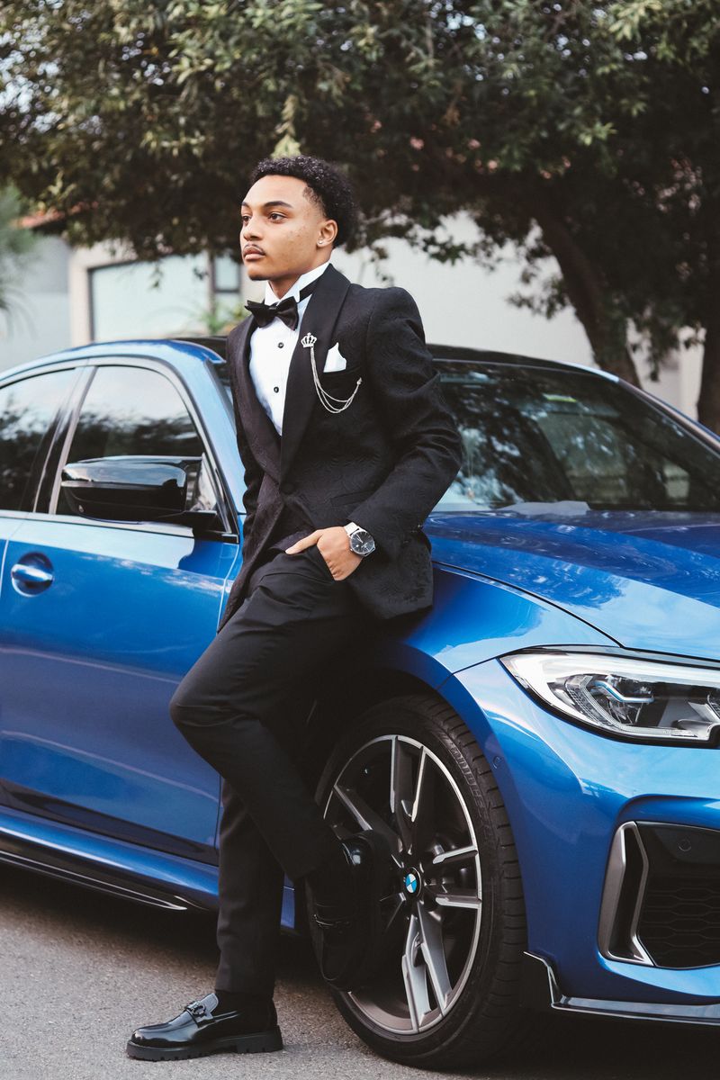 A young man in a black tuxedo leaning against a blue BMW