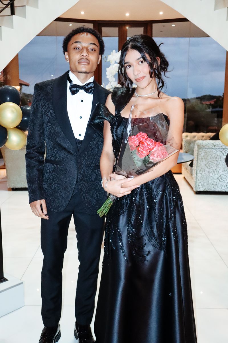 A couple dressed for a matric dance indoors, holding a bouquet of roses