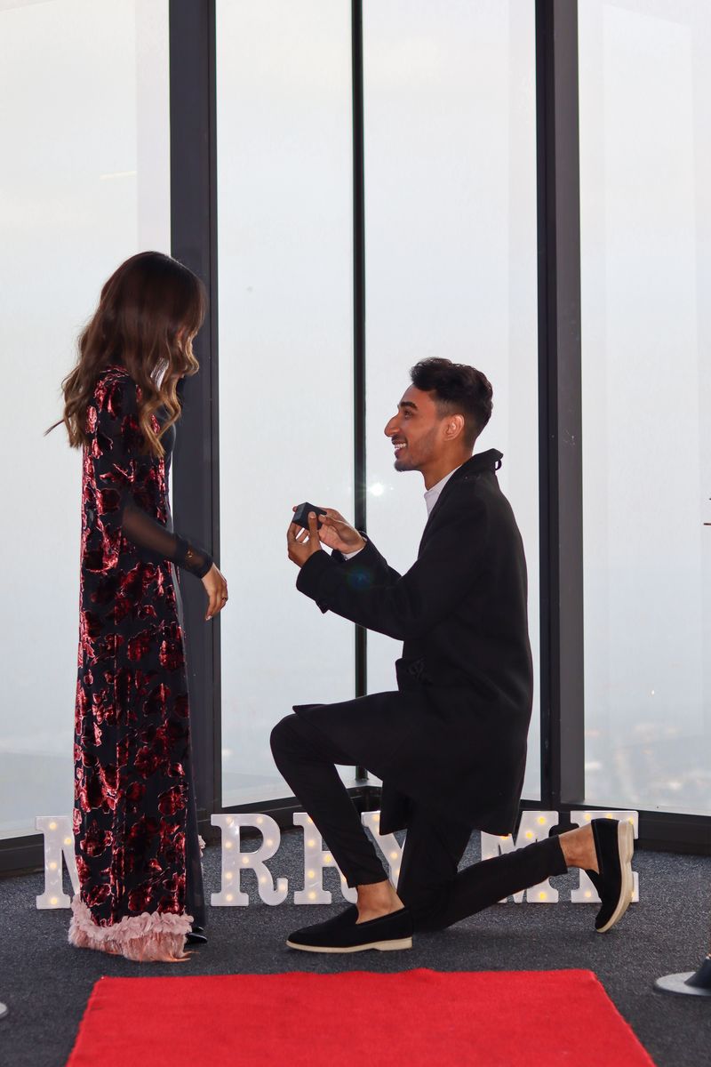 A man on one knee holding out a ring in front of illuminated MARRY ME letters
