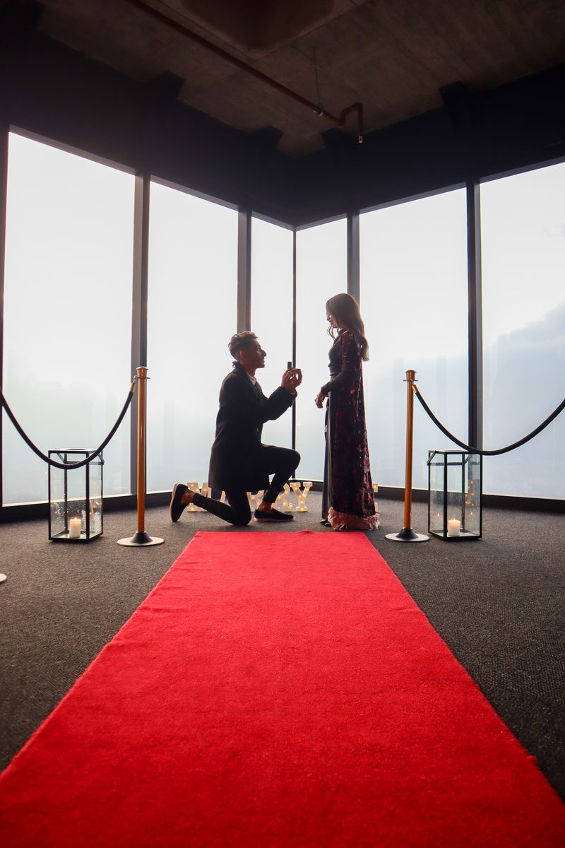 A proposal set up on a red carpet with lanterns in a room with floor to ceiling windows