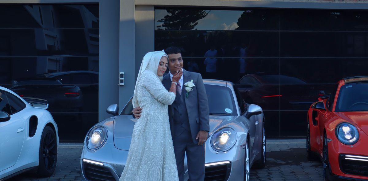 A bride and groom embracing between two parked Porsches under a building canopy